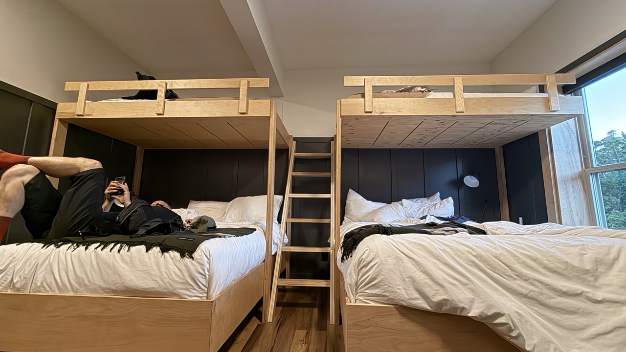 Interior view of a cozy accommodation featuring two sets of bunk beds, one on each side. A person is lounging on the lower bed, using a phone, while the upper bed is made up. The decor includes wooden structures and a large window allowing natural light to enter.