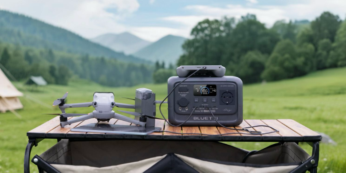 A BLUETTI Elite 30 V2 power station is set on a wooden table outdoors, beside a drone, against a backdrop of lush green mountains and clear skies.