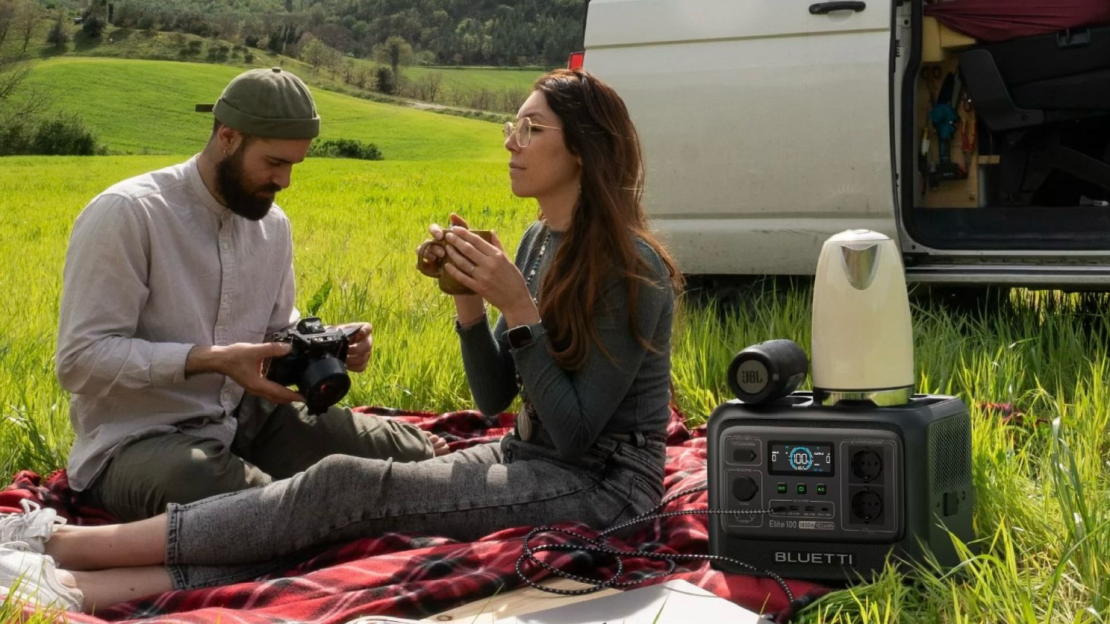 A man and a woman sit on a picnic blanket in a grassy field, with a BLUETTI portable power station in the foreground. The man is holding a camera, while the woman is drinking from a cup, enjoying the outdoor setting.
