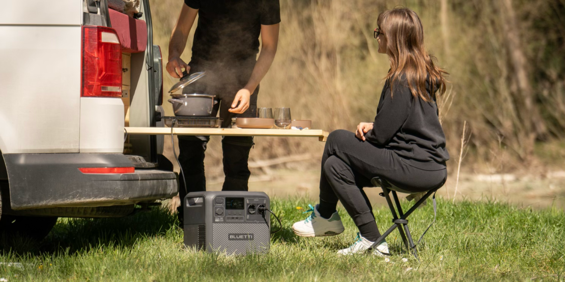 A woman sitting on a folding chair next to a portable power station while a man cooks outdoors near a van.