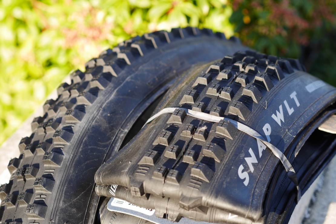 Close-up of two mountain bike tires, labeled 'SNAP WLT', bundled together with a rubber band. The tires feature aggressive tread patterns, resting against a natural backdrop of green foliage.