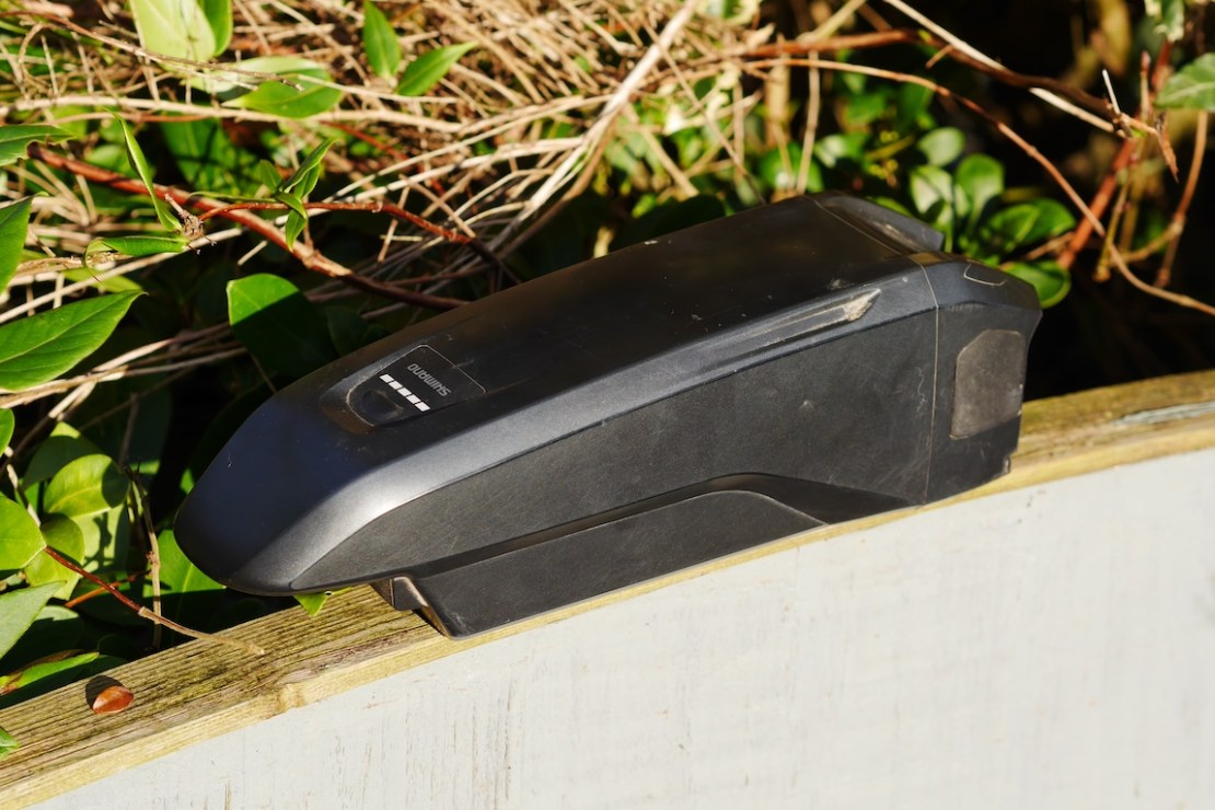 A Shimano battery resting on a wooden surface surrounded by greenery.