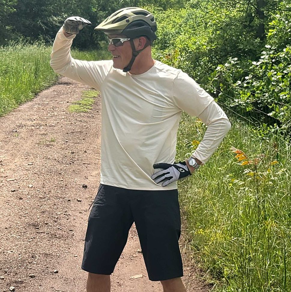 Chipps wearing a helmet and sunglasses stands on a dirt path, shielding his eyes while looking into the distance among tall green grass and trees.