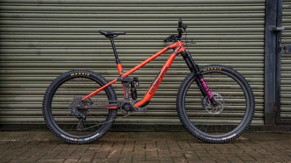 A side view of a mountain bike with a sleek carbon frame in orange and black colors, positioned against a textured green background.