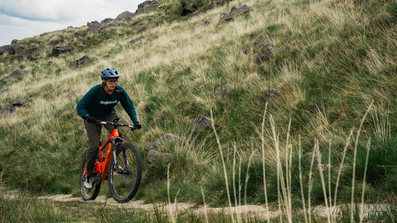 A cyclist riding an orange mountain bike on a grassy trail amidst rocky terrain, wearing a helmet and Singletrack branded clothing.