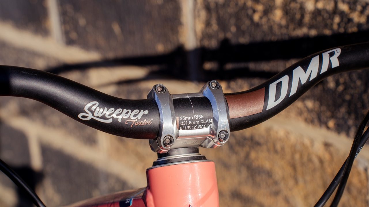 Close-up of DMR Sweeper Twelve handlebars featuring a unique back sweep angle and a colorful stem, set against a textured stone wall.