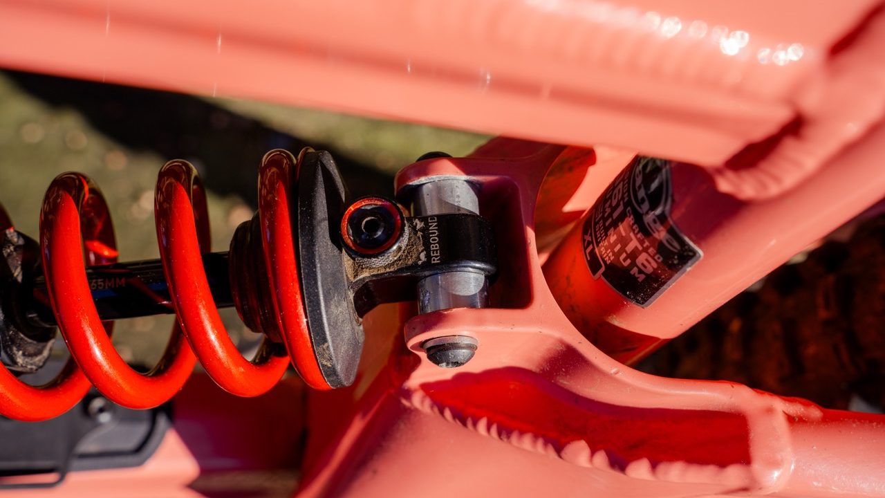 Close-up image of a mountain bike's rear suspension featuring an orange coil spring and shock absorber mounted on a pink frame.