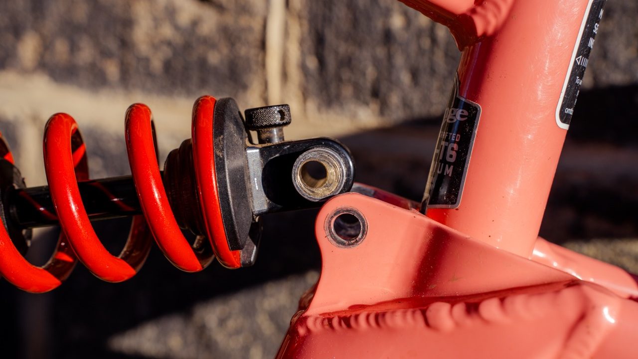 Close-up of a mountain bike's rear shock and linkage featuring a red coil spring with a black adjustment knob, mounted to a coral-colored frame.