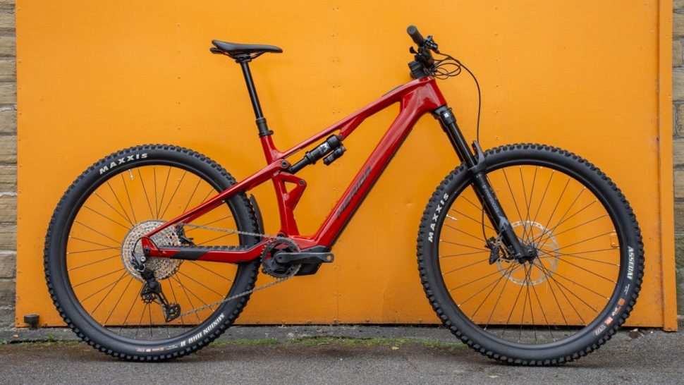 A red mountain bike displayed against an orange background, showcasing its suspension, tires, and overall design.