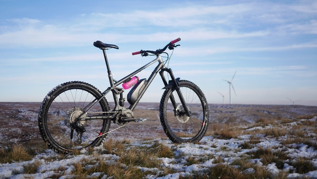 A mountain bike with a sleek frame and pink accents rests on a snowy landscape, with wind turbines visible in the background and a clear blue sky above.