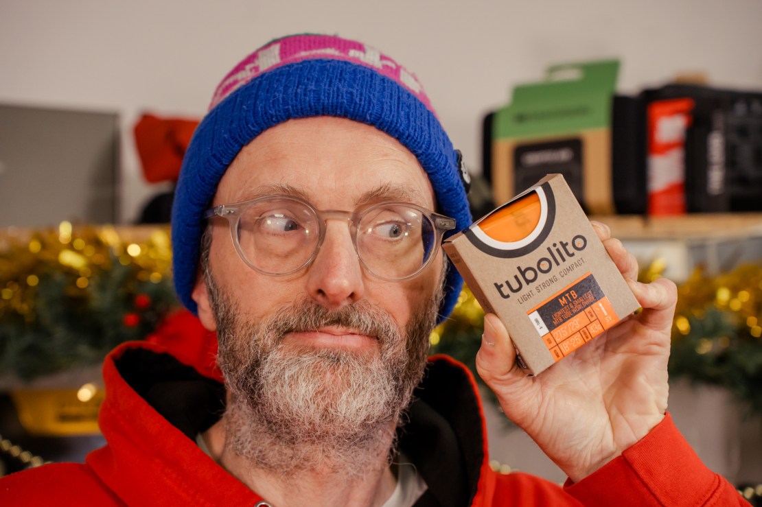 Person holding a Tubolito tube packaging with a playful expression, wearing a colorful knitted hat and red hoodie, set against a festive background.