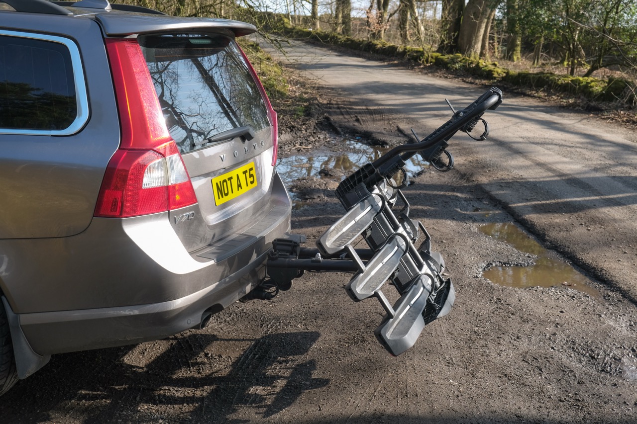 A rear view of a silver Volvo V70 with a bike rack attached, displaying multiple bike holder arms, set against a gravel road and wooded background.