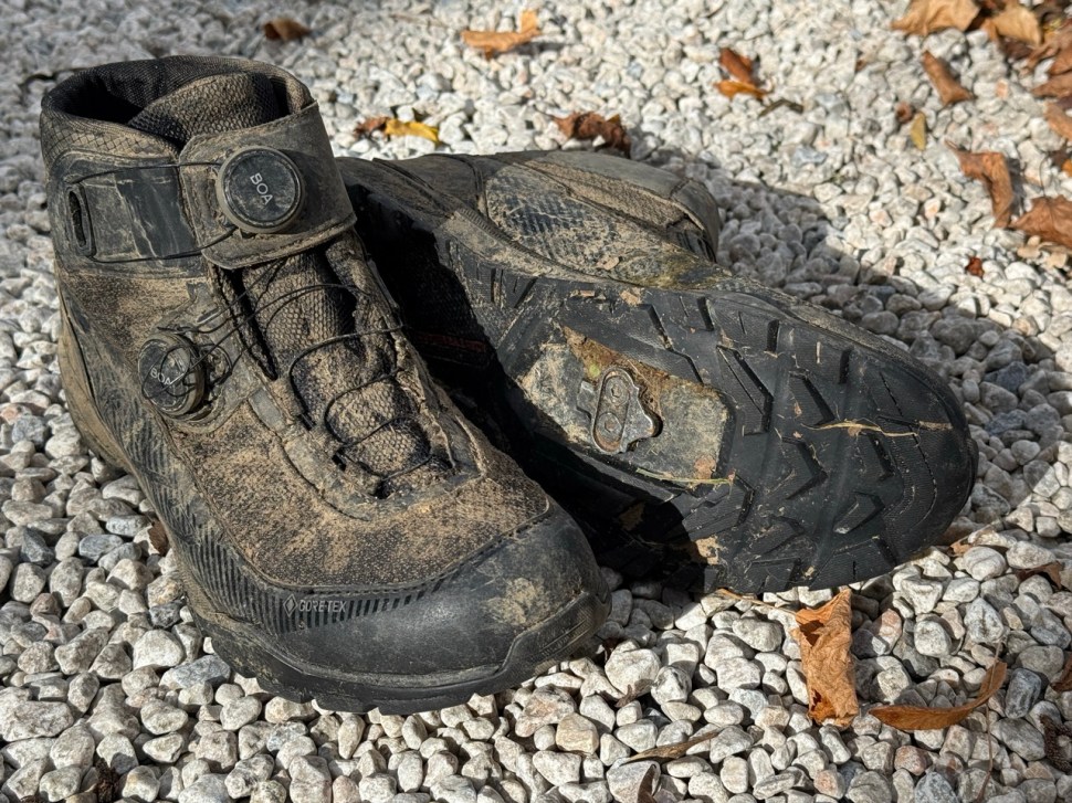 A pair of muddy Shimano EX9 winter cycling boots featuring BOA dials for a secure fit, placed on gravel with autumn leaves scattered around.