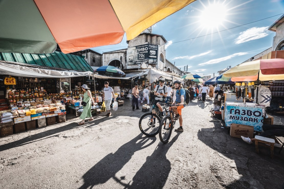 A vibrant scene at a market in Bishkek, Kyrgyzstan, with colorful umbrellas overhead, showcasing various food stalls. Two mountain bikers pause with their bikes, surrounded by bustling shoppers and a sunlit sky.
