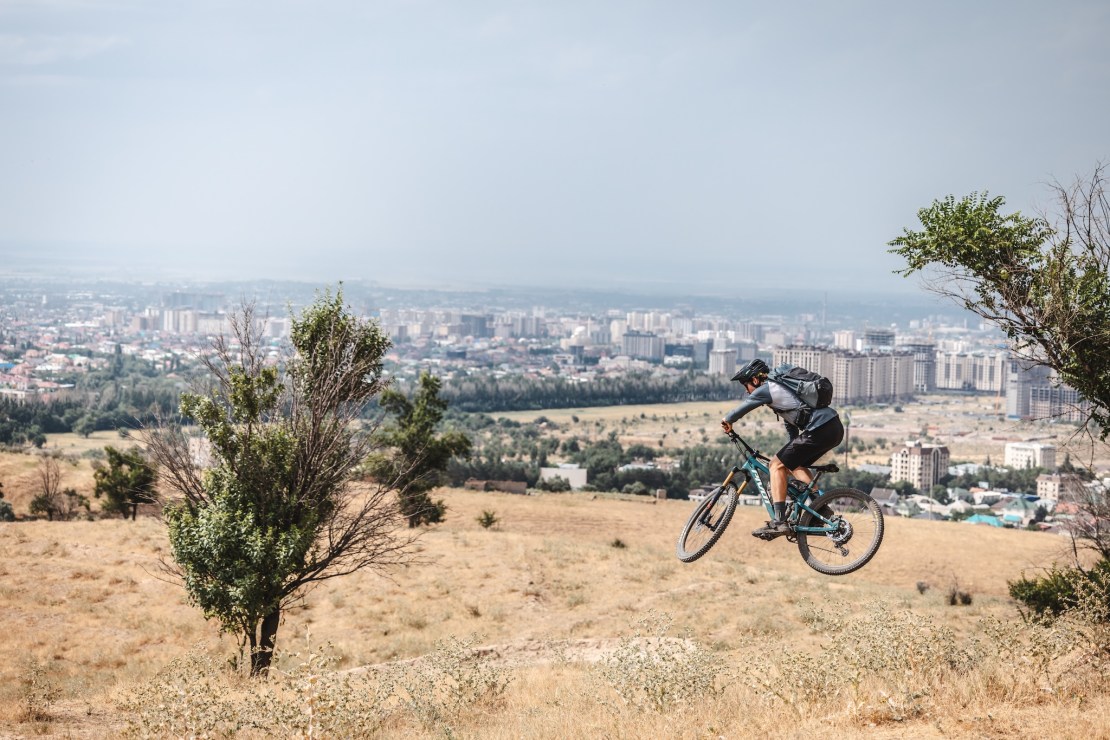 A mountain biker is jumping off a hill with a city skyline in the background, surrounded by dry grass and a few trees.