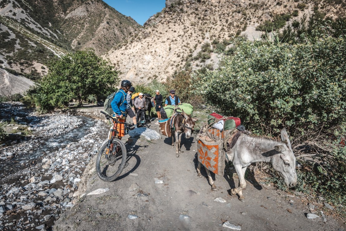 A cyclist pauses on a dirt path near a river, observing a group of people and donkeys carrying loads, set against a mountainous backdrop.