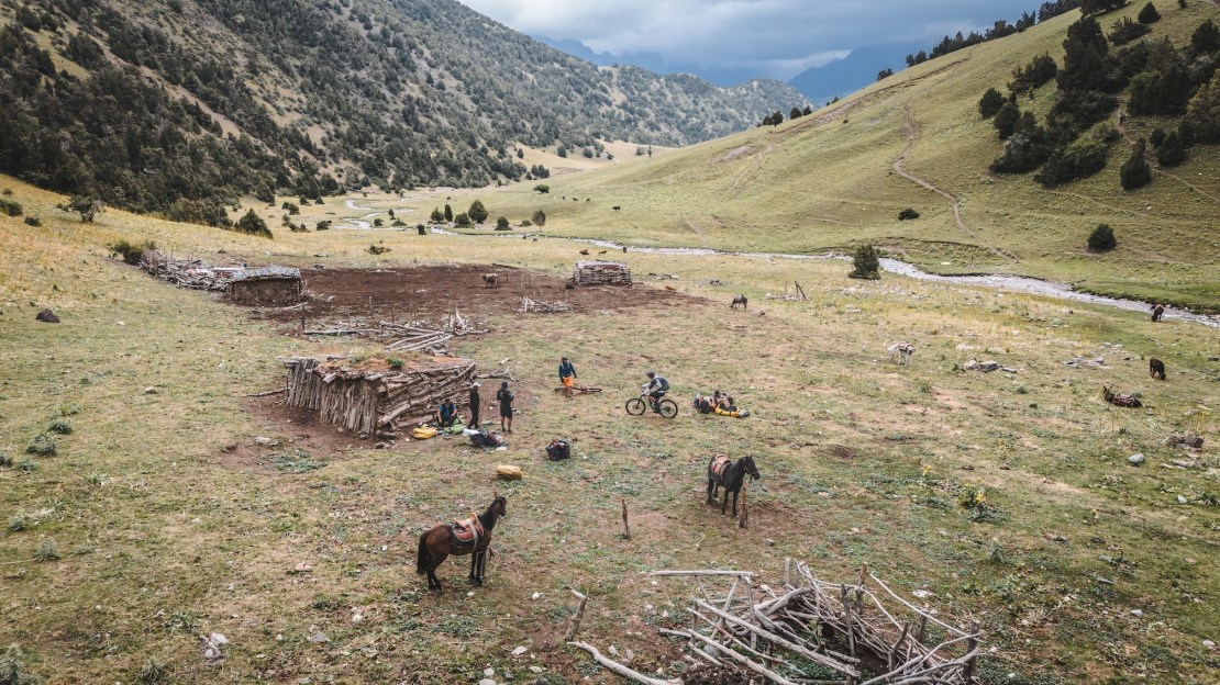 A panoramic view of a grassy valley in Kyrgyzstan featuring a rustic wooden shelter, several horses, and a group of adventurers preparing their gear in the foreground.