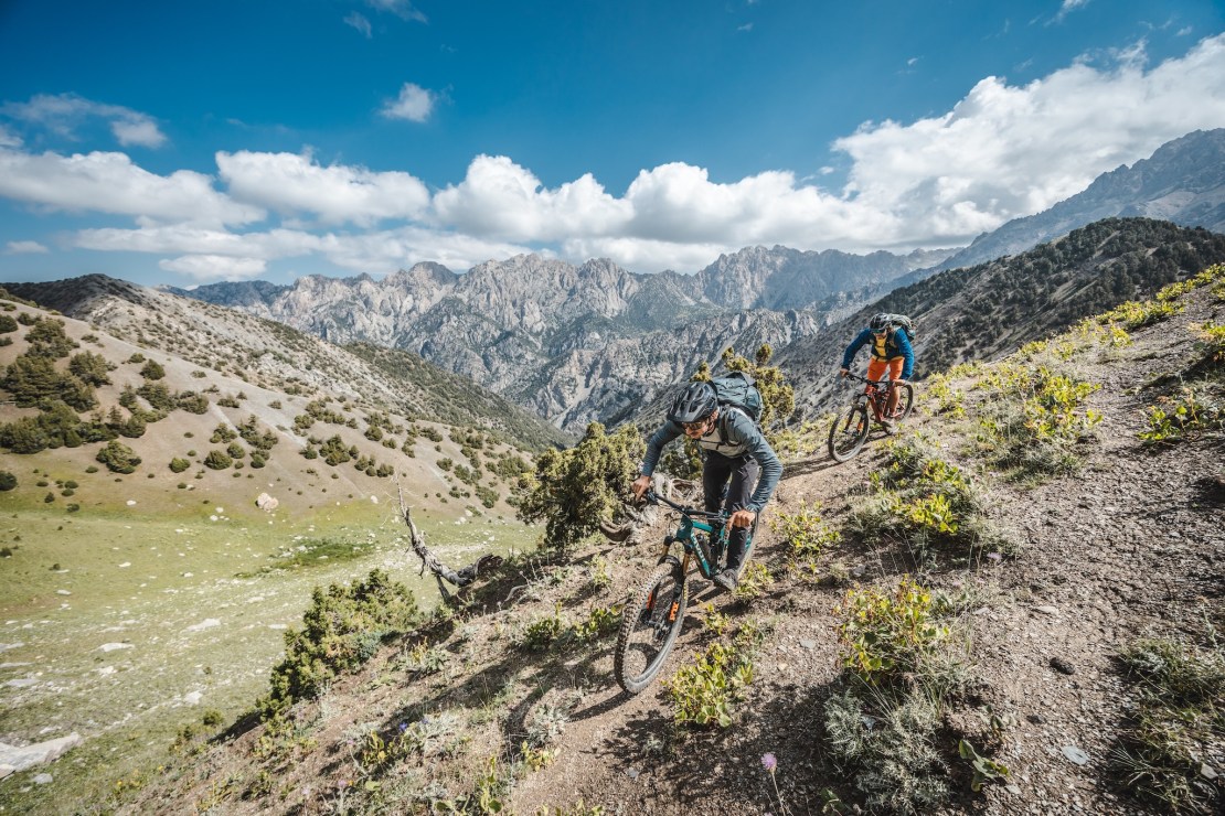 Two mountain bikers navigating a rugged trail in the Pamir Mountains, with steep rocky peaks and greenery in the background under a partly cloudy sky.