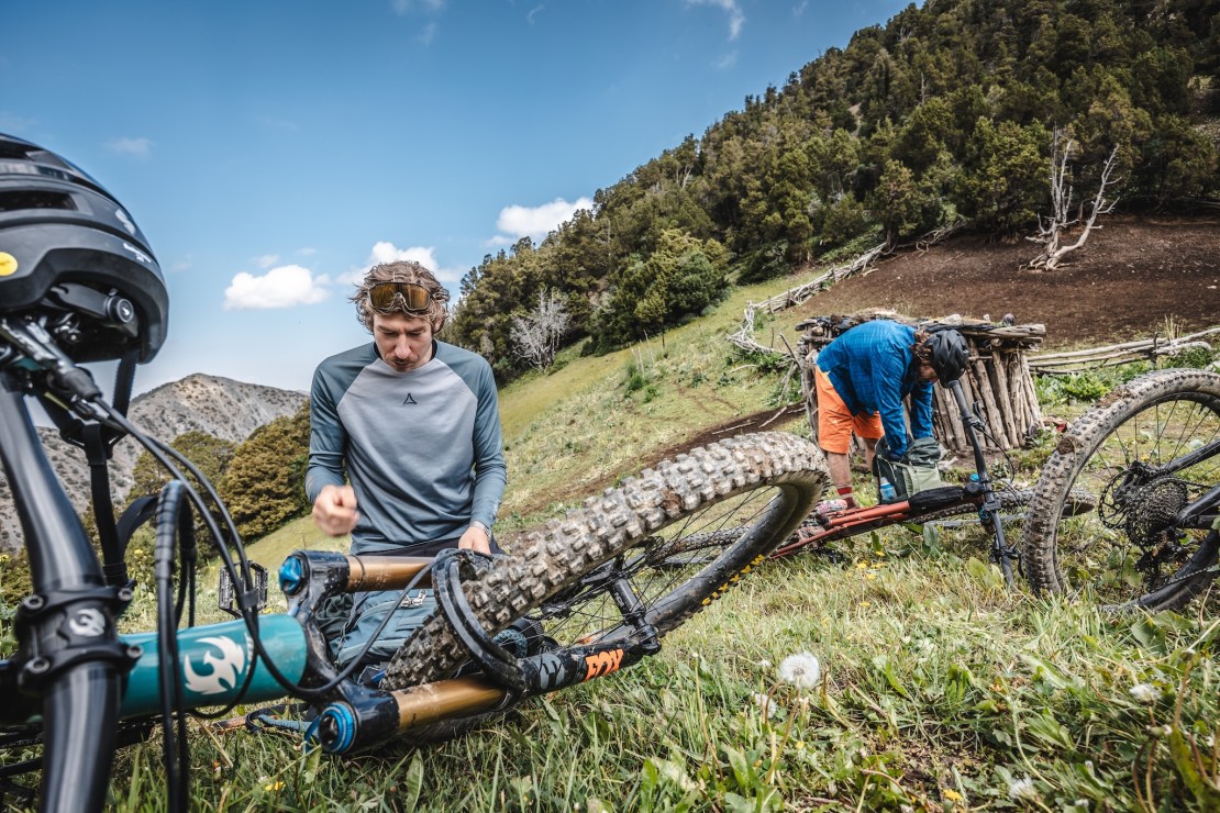 Two mountain bikers working on their bikes in a grassy area surrounded by hills and trees.