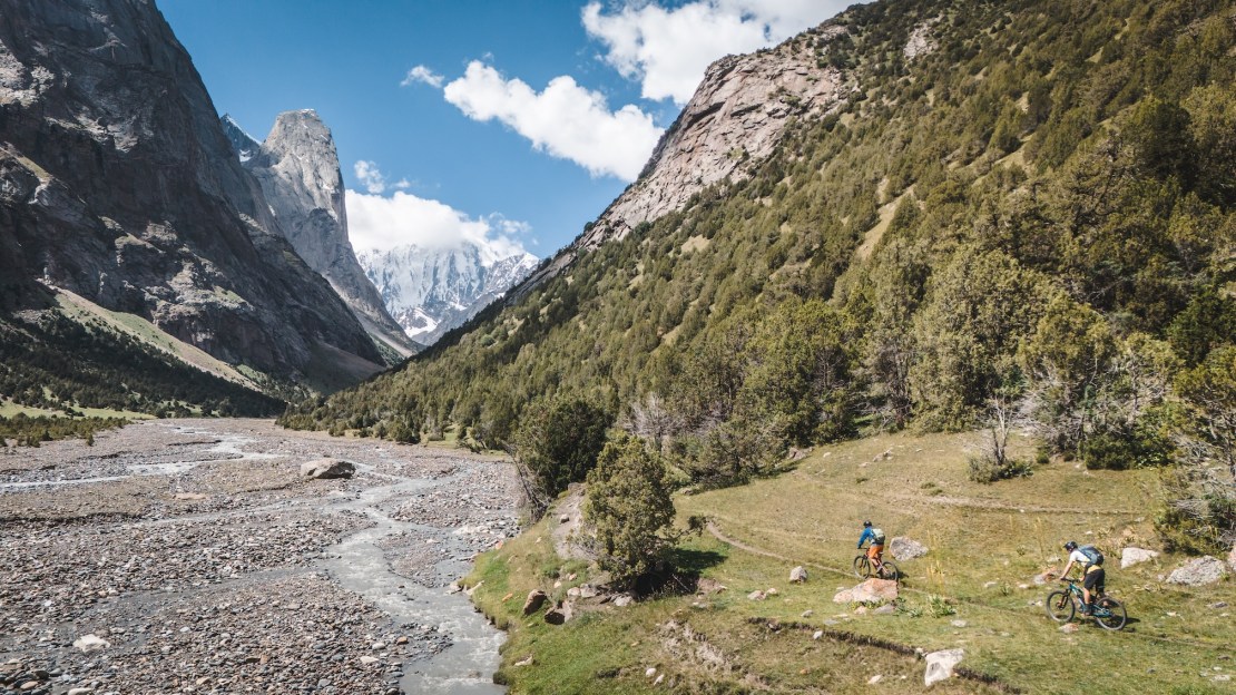 Two mountain bikers navigating a rocky trail along a river in a lush valley, surrounded by towering mountains under a blue sky with fluffy clouds.