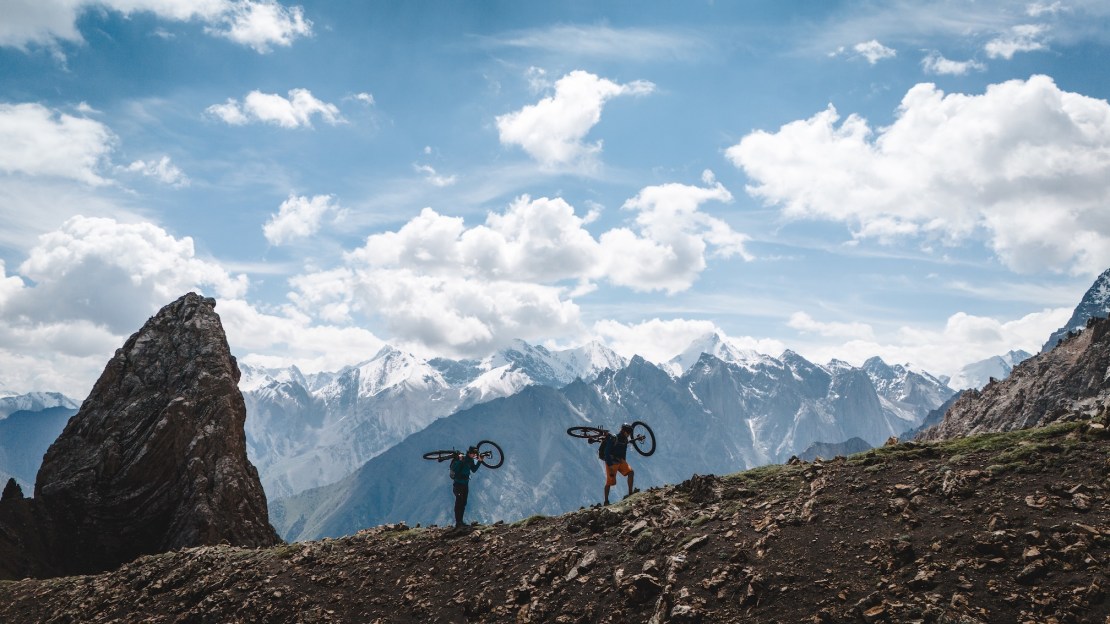 Two mountain bikers carry their bikes while hiking along a rocky path with dramatic snow-capped peaks in the background under a cloudy sky.