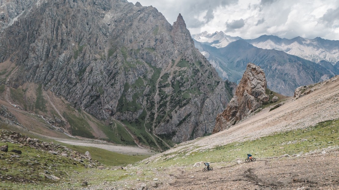 Two mountain bikers riding through a rugged valley in the Pamir Mountains, with steep rocky peaks and a cloudy sky in the background.