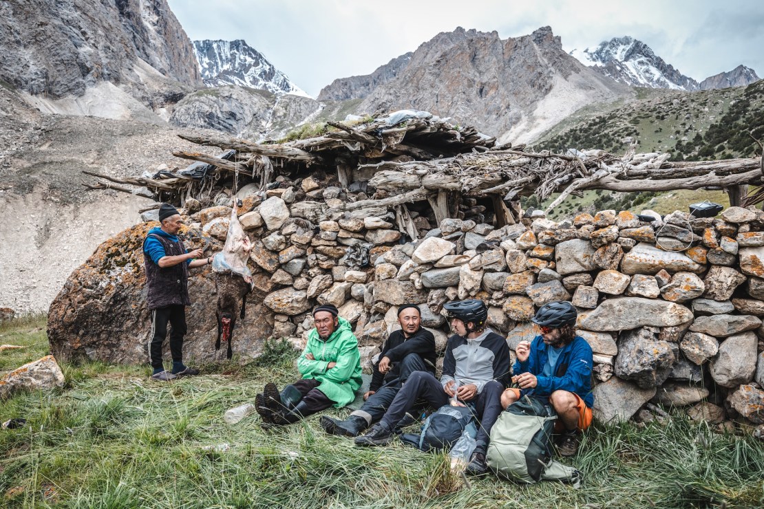 A group of cyclists resting in front of a stone shelter in the mountains, with one person engaged in preparing meat and breathtaking mountain scenery in the background.
