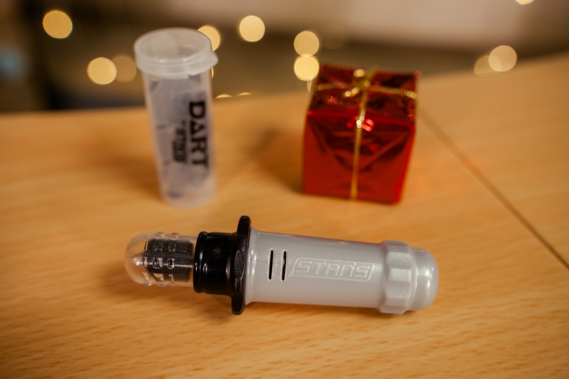 A Stan's Dart tool and refill pack displayed on a wooden surface, next to a small red gift box and a container labeled 'DART'.