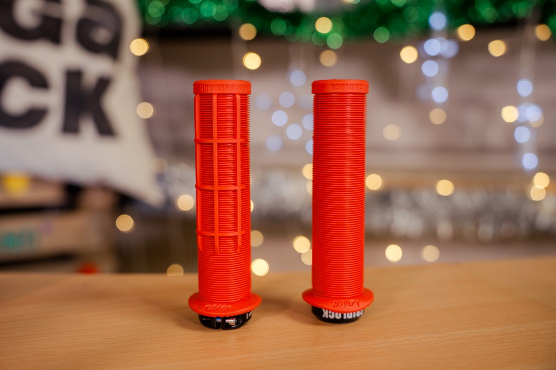 Two pairs of DMR Gridlock lock-on grips in vibrant red color, displayed on a wooden surface with blurred festive lights in the background.