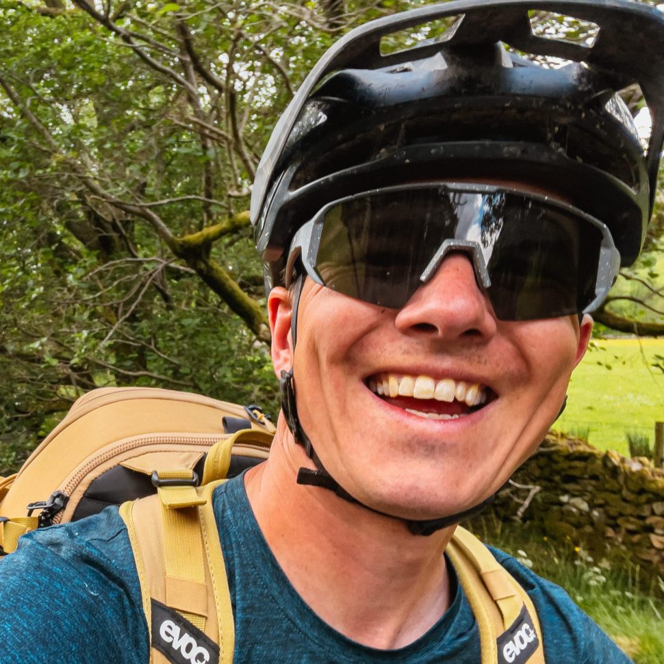 A smiling individual wearing a black helmet and sunglasses, with a backpack, cycling outdoors among greenery.