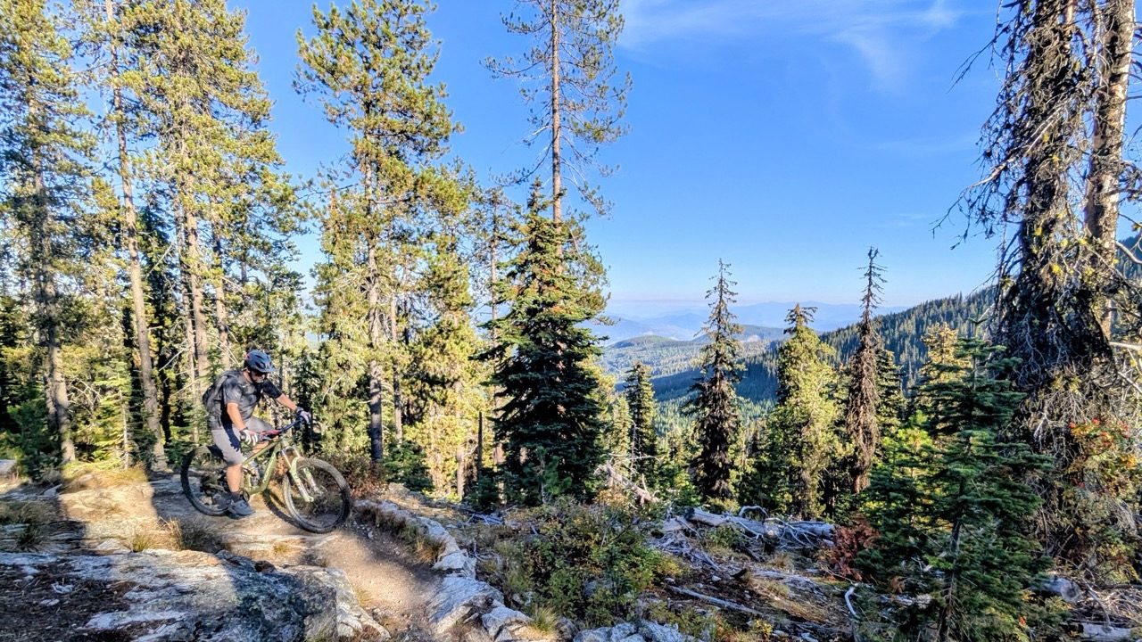 A mountain biker navigating a rocky trail surrounded by tall trees and scenic mountain views in Rossland, British Columbia.