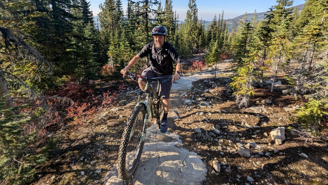 A mountain biker navigating a rocky trail surrounded by tall trees in the Kootenay region of British Columbia on a sunny day.