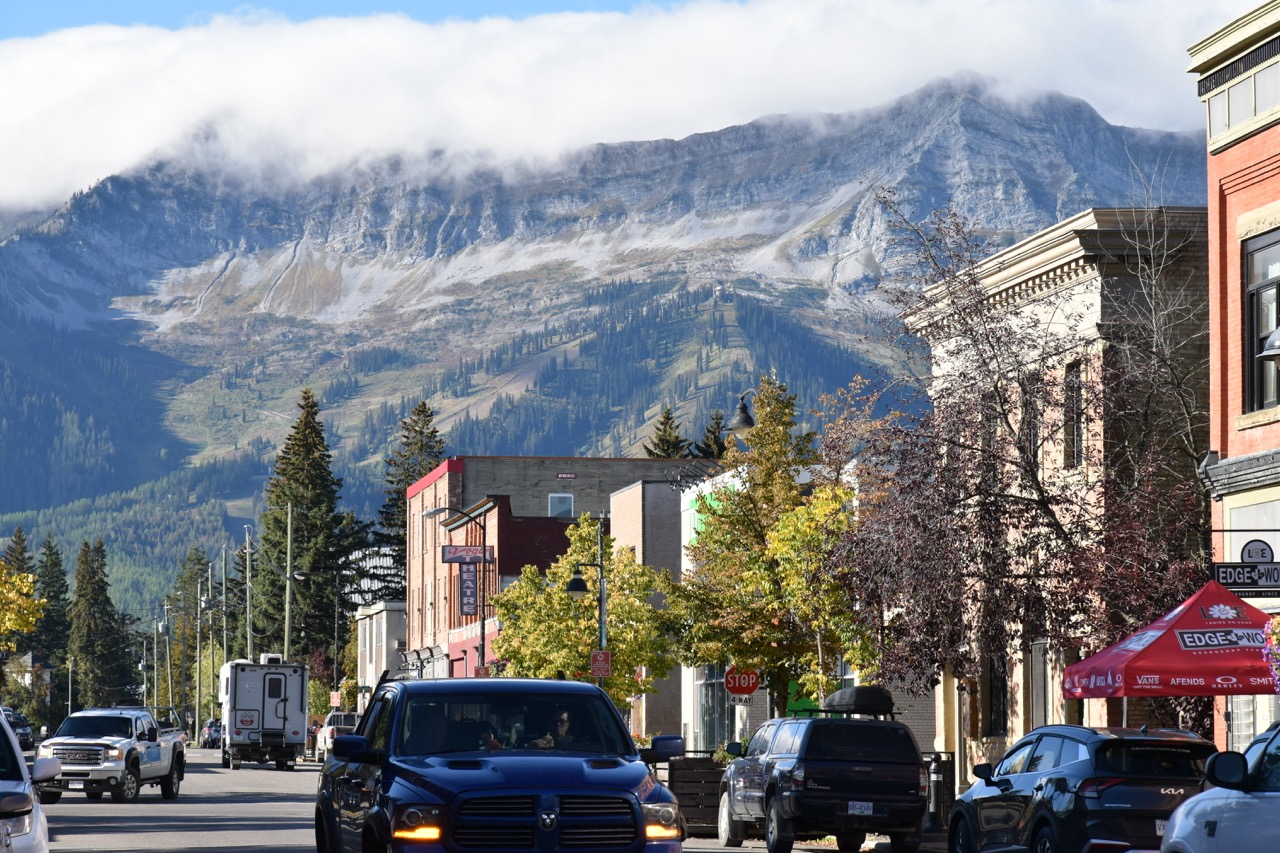A street view of Fernie, Canada, featuring a busy road lined with cars, shops, and trees, set against a backdrop of the Rocky Mountains shrouded in clouds.