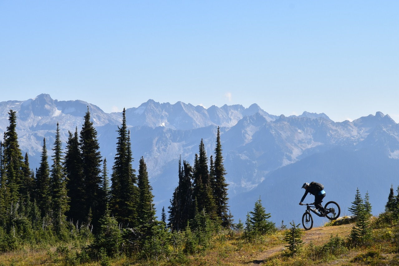A mountain biker navigating a rocky trail with tall evergreen trees and a panoramic view of snow-capped mountains in the background.