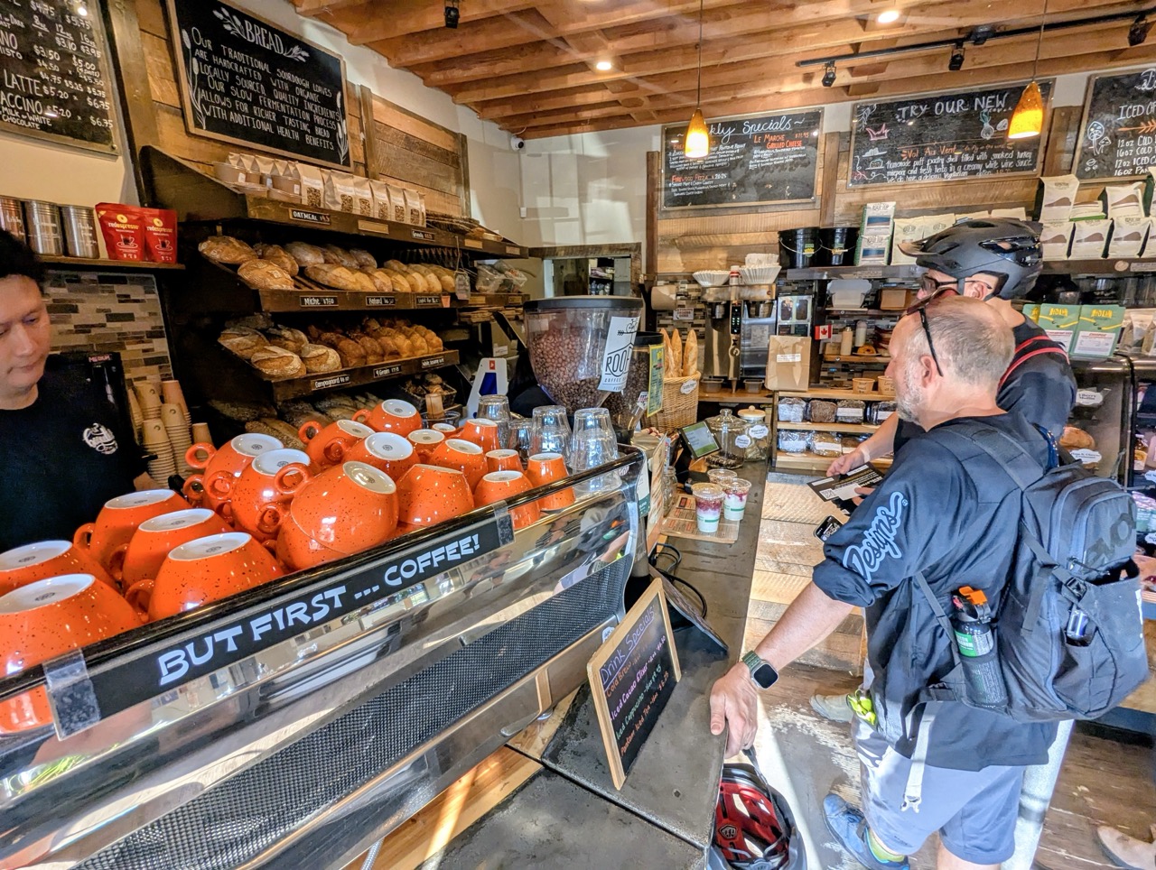 Two cyclists ordering coffee at a cozy cafรฉ with shelves of baked goods in the background.