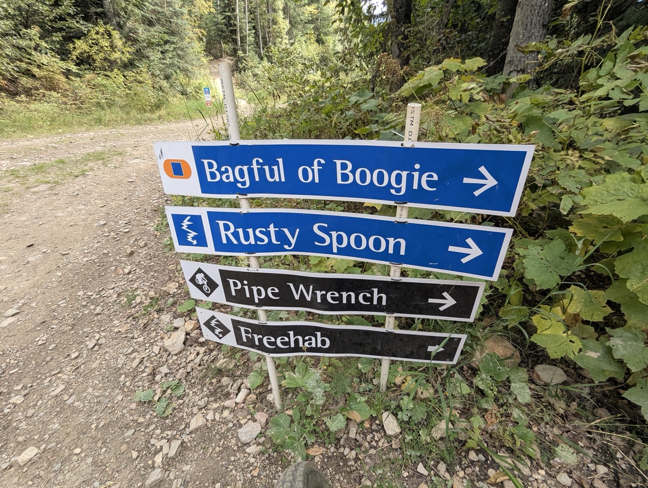 Trail signpost displaying bike trail names: Bagful of Boogie, Rusty Spoon, Pipe Wrench, and Freehab, surrounded by greenery.