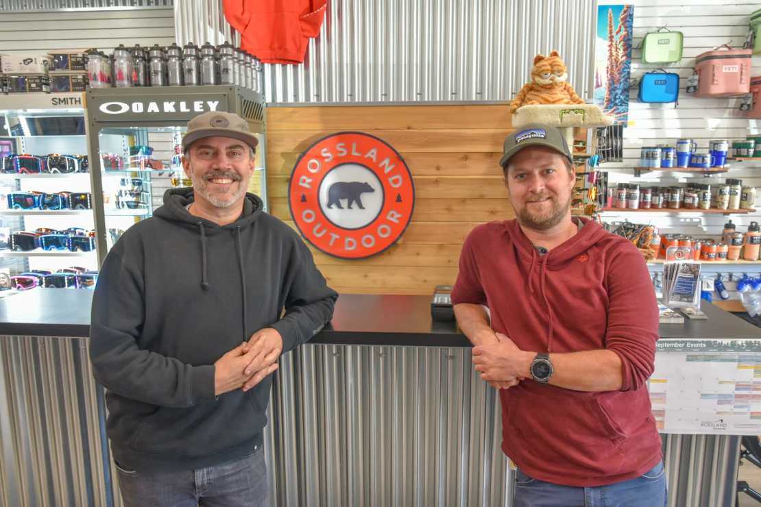 Two men smiling behind a counter in an outdoor gear shop, with a logo of Rossland Outdoor in the background and various equipment displayed on shelves.