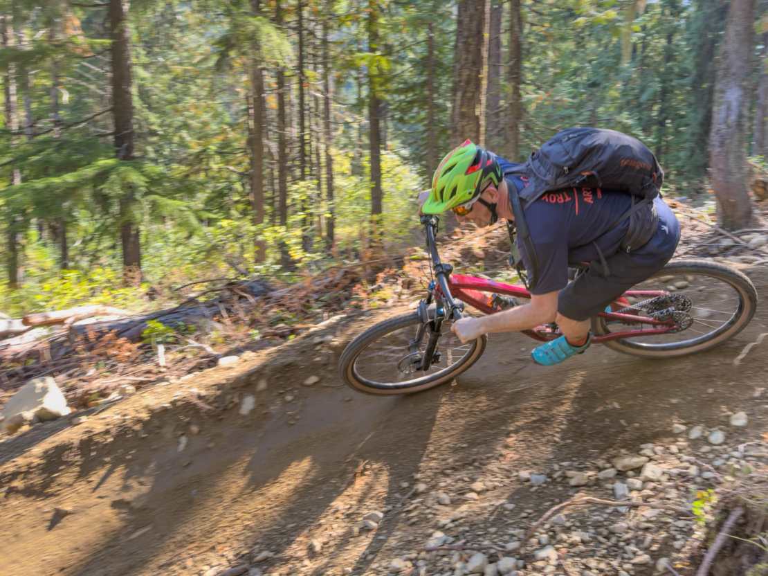 A mountain biker navigating a winding dirt trail in a forested area, surrounded by tall trees and sunlight filtering through the leaves.