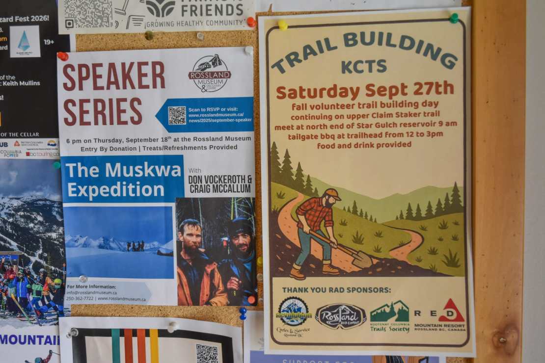 A bulletin board filled with colorful flyers, including an event announcement for a speaker series at the Rossland Museum and a call for volunteers for a trail building day organized by the Kootenay Columbia Trails Society.