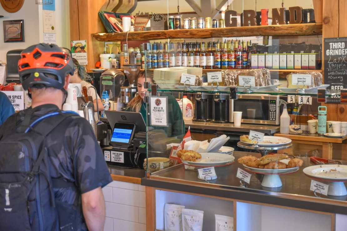 A customer in a biking outfit is waiting at the counter of a cozy café, with a barista behind the glass preparing orders. The café features a variety of coffee drinks and baked goods on display.