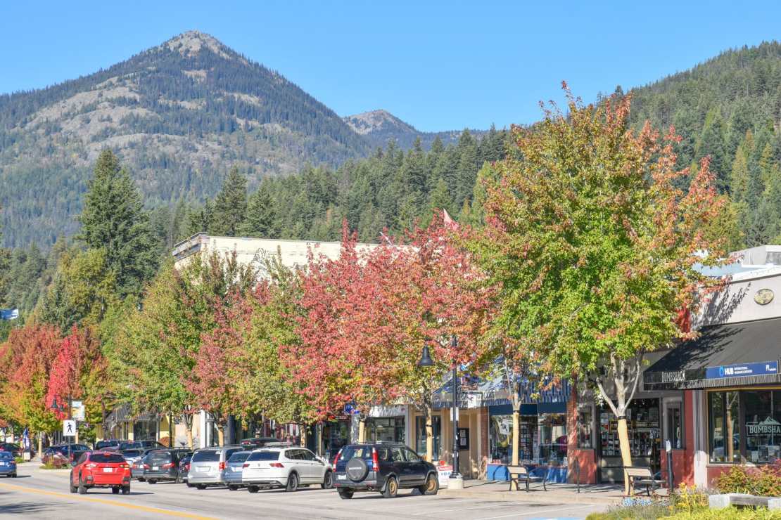 A scenic view of downtown Rossland, British Columbia, with colorful autumn trees lining the street, parked cars, and the backdrop of mountains under a clear blue sky.
