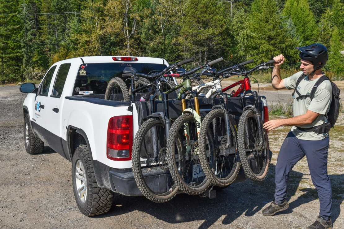 A man in a helmet is preparing to unload a mountain bike from the back of a pick-up truck, which is transporting several bikes for a ride in the forested area.