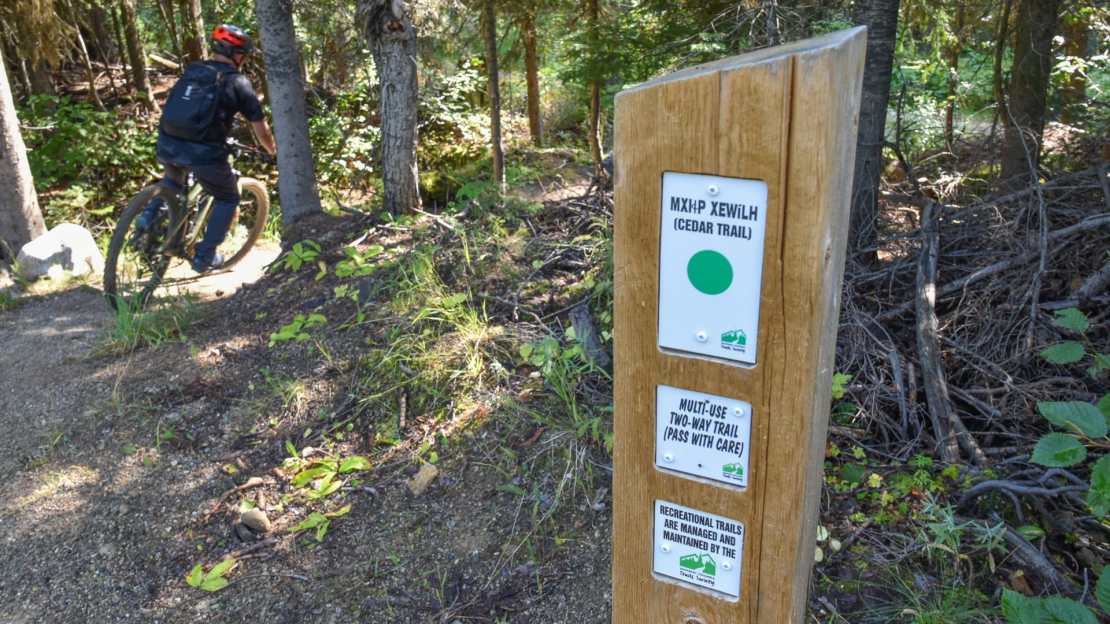 A mountain biker riding along the Cedar Trail in Rossland, British Columbia, with a trail sign indicating it's a multi-use two-way trail.