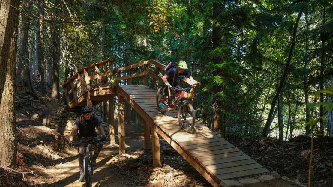 Two mountain bikers riding over a wooden bridge on a forest trail in Rossland, British Columbia, surrounded by tall trees.
