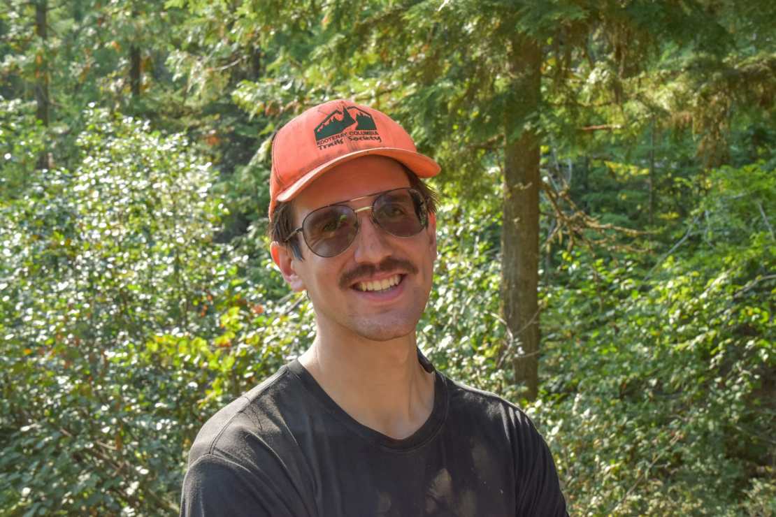 A smiling man wearing sunglasses and an orange cap stands against a backdrop of green trees, suggesting a forested outdoor setting.