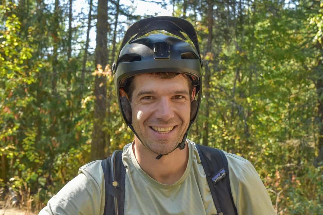 A cheerful person wearing a biking helmet and gear, smiling in a forest setting with greenery in the background.