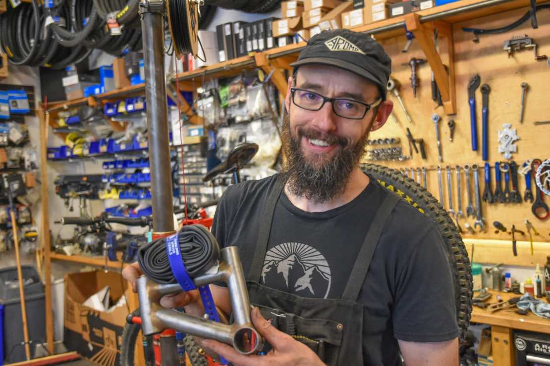 A smiling man with a beard holds a bicycle part and a rolled tire, standing in a bike shop filled with tools and bicycle accessories.