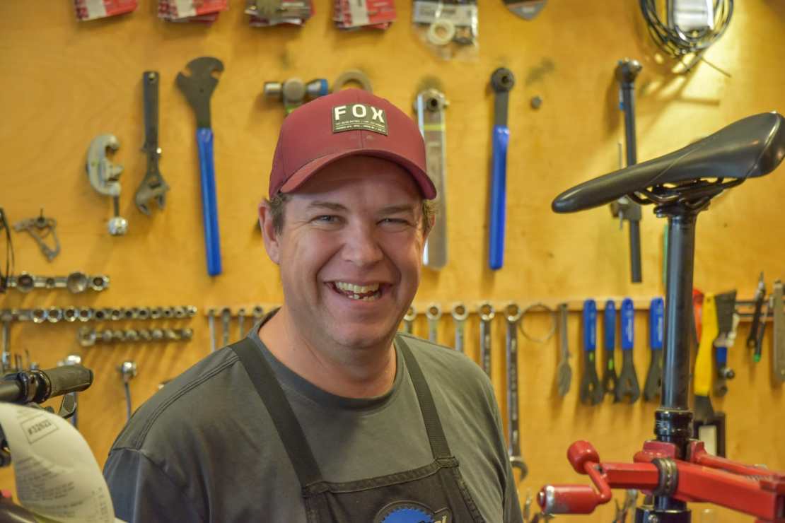 A smiling man wearing a red cap and an apron stands in a bike shop surrounded by tools and bike parts on a wooden wall.
