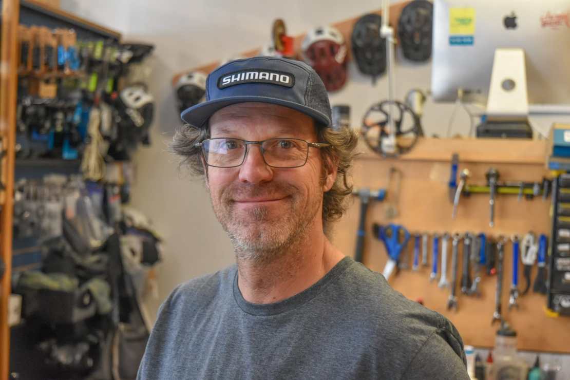 A smiling person wearing a Shimano cap and glasses is standing in a bike shop, with various bicycle tools and accessories displayed in the background.