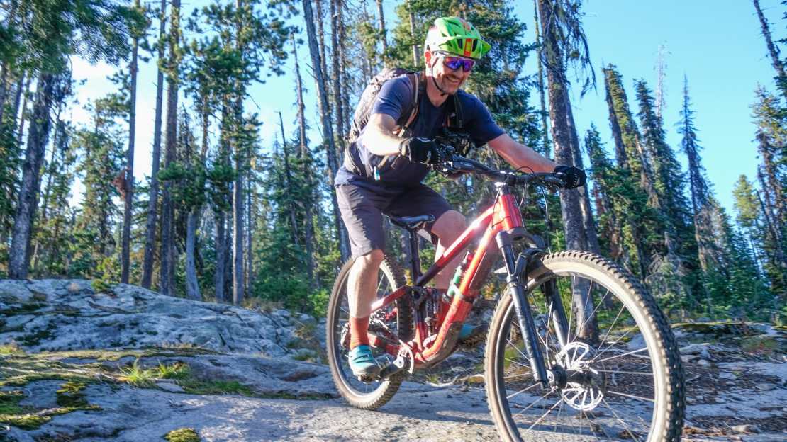 A mountain biker navigating a rocky trail surrounded by tall coniferous trees.